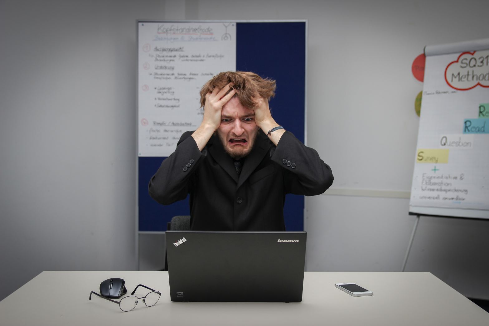 30-something white man in black business clothes with hands to head and upset look on face in front of laptop