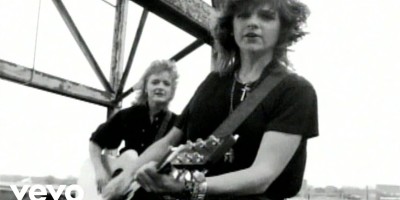 Amy Ray and Emily Saliers, the Indigo Girls, in black/white photo playing guitars in front of industrial structure