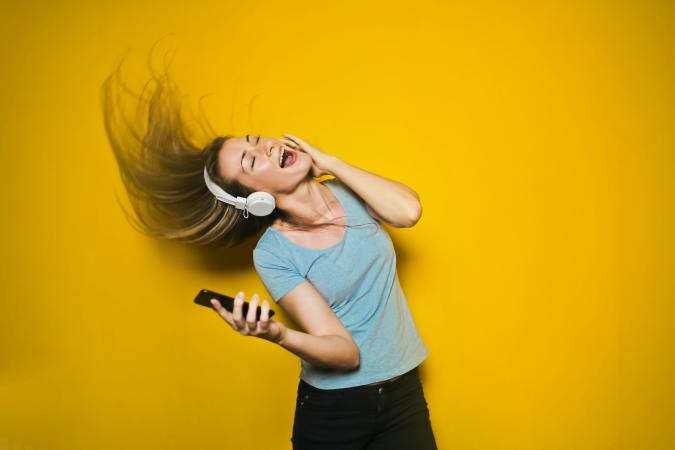 woman with long brown hair swinging head with white headphones on, holding smart phone, and singing